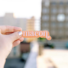 Load image into Gallery viewer, Up close photo of a white woman&#39;s hand holding a sticker over the Macon skyline. The sticker says &quot;Macon&quot; in light pink in a lower case serif font with ball flourishes on the &quot;m&quot; and the descender of the &quot;n.&quot; The word has a angled drop shadow in orange.
