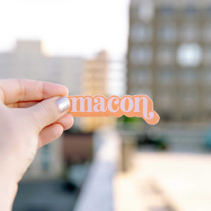 Up close photo of a white woman's hand holding a sticker over the Macon skyline. The sticker says "Macon" in light pink in a lower case serif font with ball flourishes on the "m" and the descender of the "n." The word has a angled drop shadow in orange.