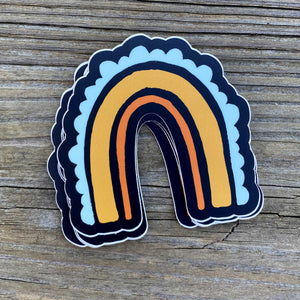 On a wooden table rests a stack of  3x3" rainbow stickers. The rainbow has three stripes. The inside one is a thin orange line, the middle one is a golden rod thicker line, and the outer stripe is a team scalloped line. The background color is black.