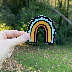 A white woman holds up a 3x3" rainbow sticker in front of greenery. The rainbow has three stripes. The inside one is a thin orange line, the middle one is a golden rod thicker line, and the outer stripe is a team scalloped line. The background color is black.