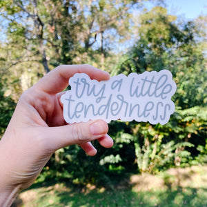 Woman's hand holding a white sticker with "try a little tenderness" hand lettering with greenery in background