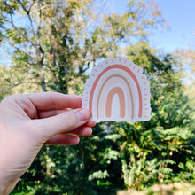 Load image into Gallery viewer, Woman&#39;s hand holding a rainbow shaped sticker with pink tones with trees in the background

