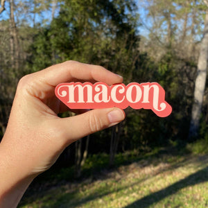 Hand holding a pink sticker with "macon" text in lighter pink color with greenery in the background