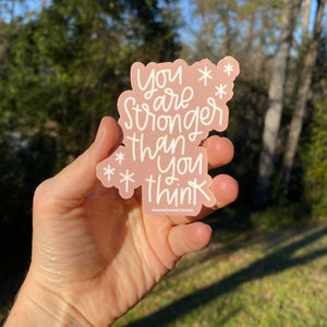 Woman's hand holding a pink sticker with white hand lettering that reads "you are stronger than you think" with trees in the background
