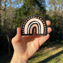 Load image into Gallery viewer, Hand holding a black sticker with a rainbow design in pink tones with greenery in the background

