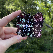 Load image into Gallery viewer, Woman holding a black sticker with colorful hologram hand lettering that reads &quot;you are made of magic&quot; surrounded by stars. The sticker is being held in front of greenery.
