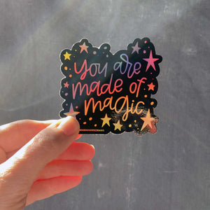 Woman holding a black sticker with colorful hologram hand lettering that reads "you are made of magic" with stars.