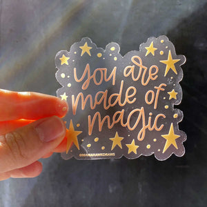 Woman holds a clear sticker with yellow stars on the border and pink hand lettering that reads "you are made of magic"