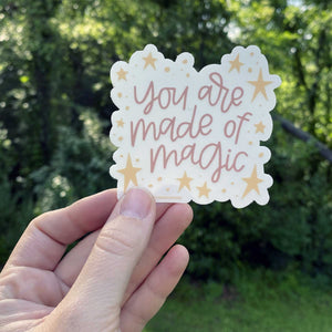 Woman holds a clear sticker with yellow stars on the border and pink hand lettering that reads "you are made of magic." The sticker is being held in front of greenery.