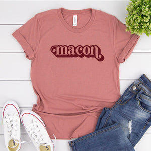 Pink tee with "macon" text in center on a white wooden background with white sneakers, jeans, and foliage in the background