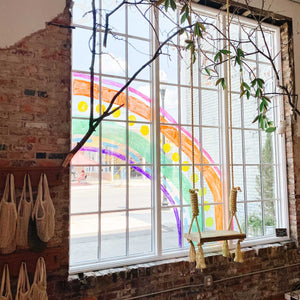 Indoor view of 6x9 window with panes with a rope swing and soft greenery in the foreground. The window has a rainbow painted with lines and shapes and rainbow colors.