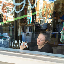Load image into Gallery viewer, Sidewalk view of Mama Hawk Draws painting a window from inside with &quot;#FRANK&quot; in white paint.
